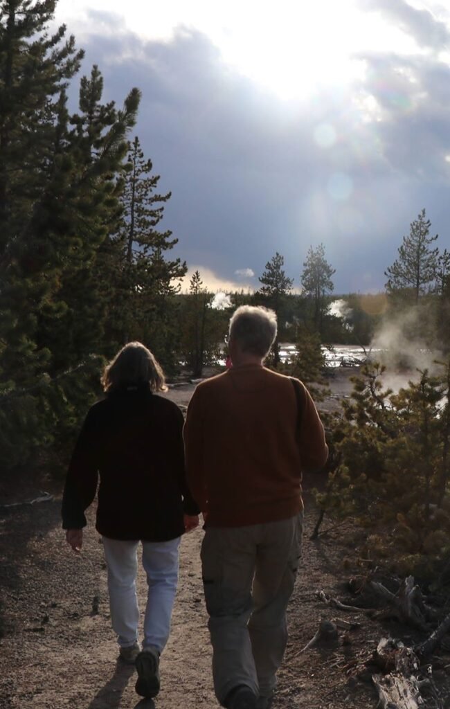 A woman and a man walking away from the photographer on a path with evergreen trees and steam from a nearby geothermal pool.