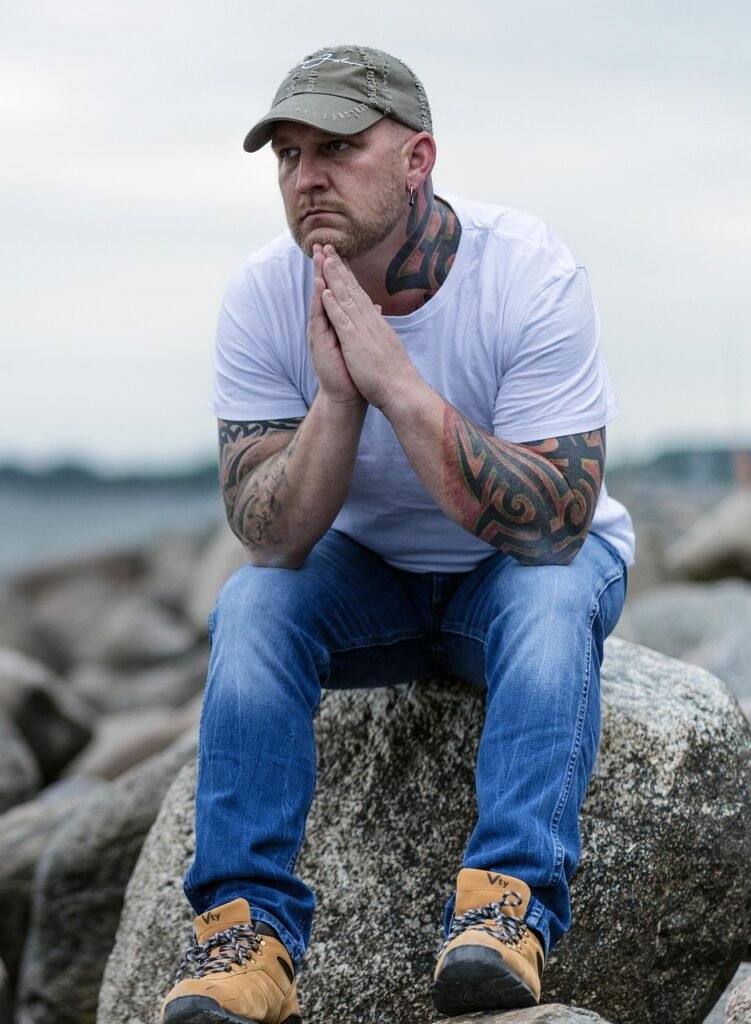 Photo of a young man in blue jeans and a baseball cap sitting on a boulder on the shore.