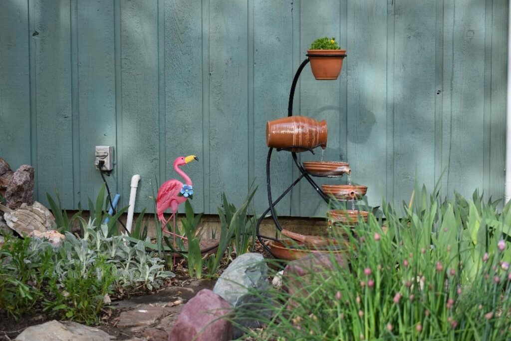 A photograph of a unique garden water fountain and a pink flamingo in a flower garden.