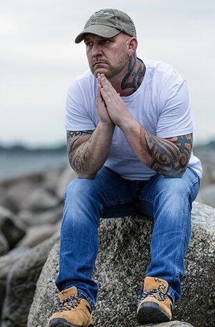 A young man with tatoos sits on a large granite boulder on the shoreline, looking thoughtfully into the distance.
